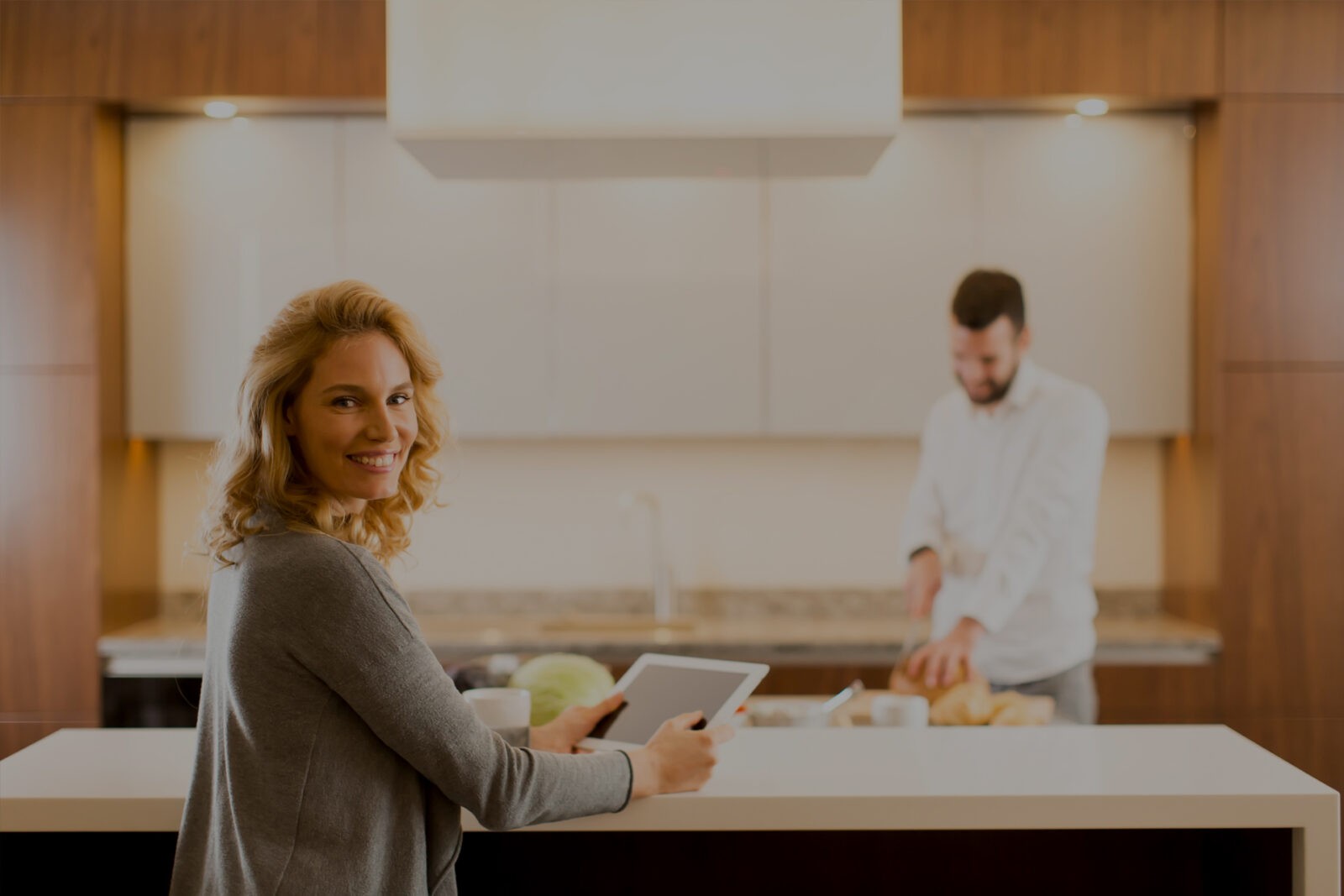 Kobieta steruje smart home tabletem w nowoczesnej kuchni, a w tle mężczyzna przygotowuje posiłek.