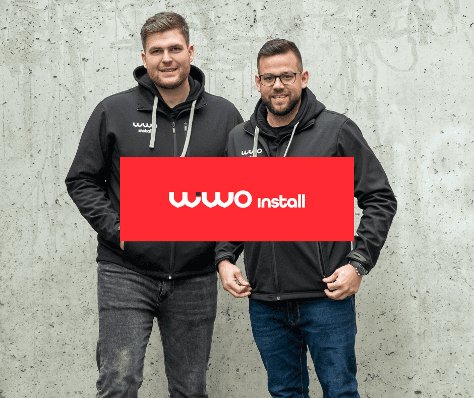 Dwóch pracowników Wiwoinstall w czarnych bluzach z logo firmy stoi przed betonową ścianą, a na środku widoczny jest czerwony baner z napisem Wiwo install.